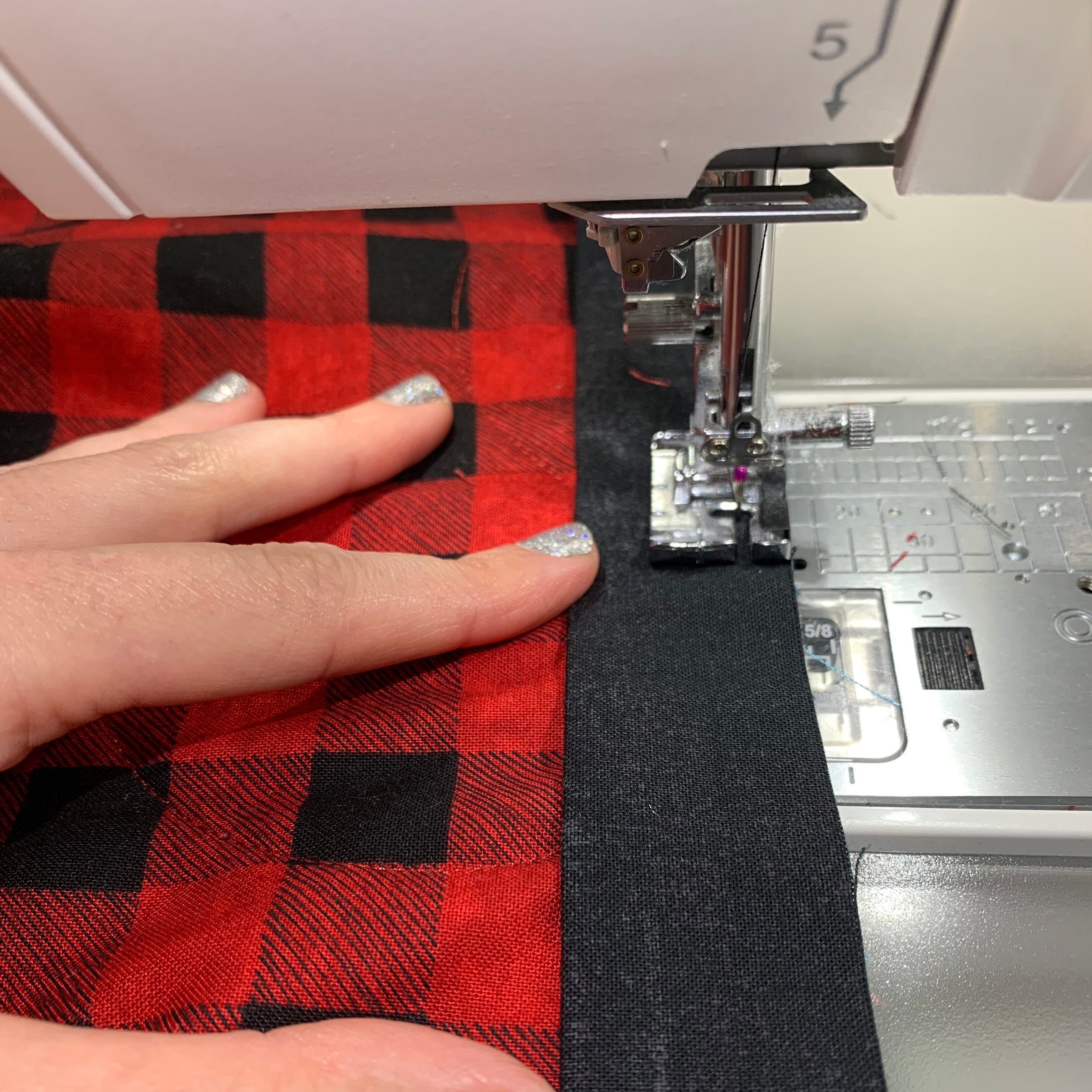 A close-up of a person's hand guiding a red and black buffalo plaid fabric through a white sewing machine.