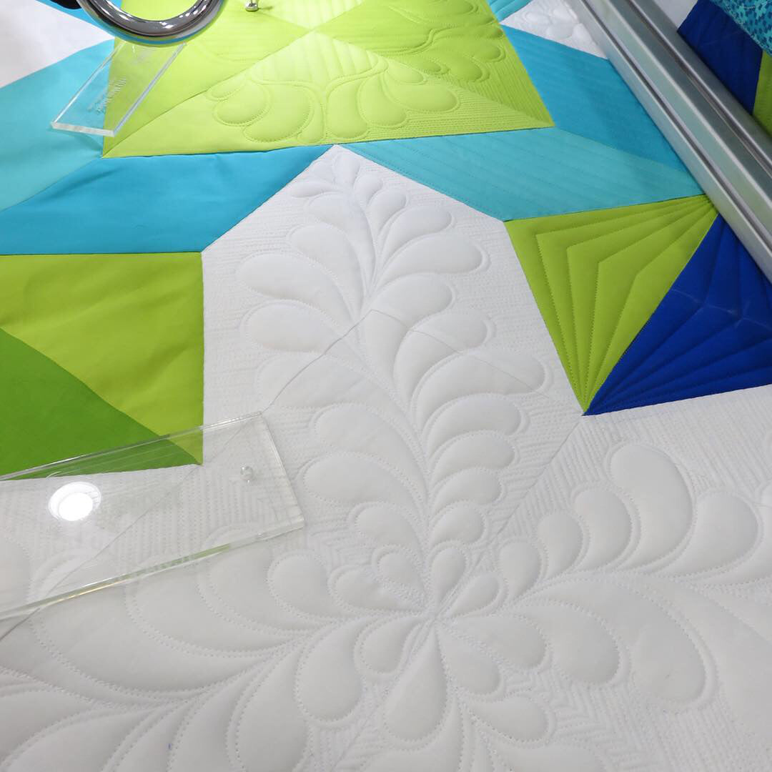 A close-up of a quilted fabric block with a white floral pattern, surrounded by geometric sections of turquoise, lime green, and blue. A quilting machine foot is visible at the top edge, and a clear acrylic guide is placed on the fabric.
