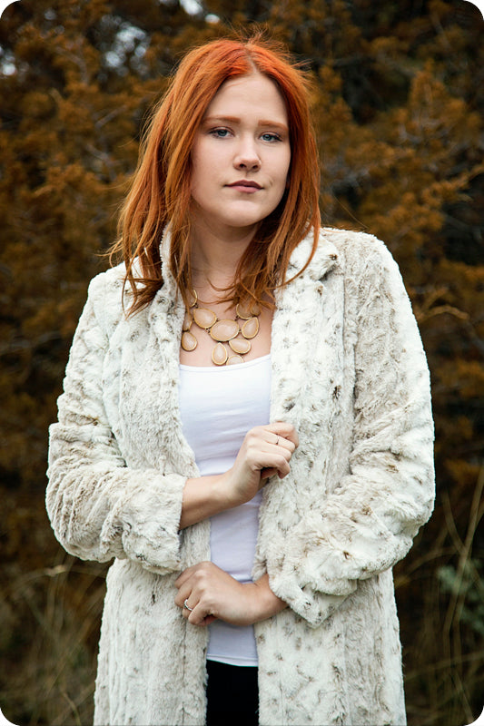A woman with red hair wearing white and standing in front of bushes.