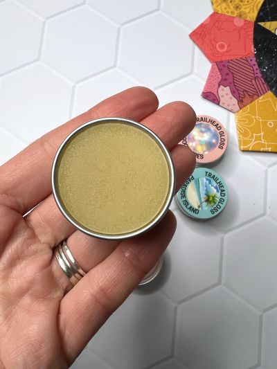 Hand holding a small tin of yellow balm with colorful tins in the background.