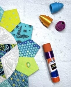 Crafting materials including glue stick, fabric hexagons, and sewing tools on a table.
